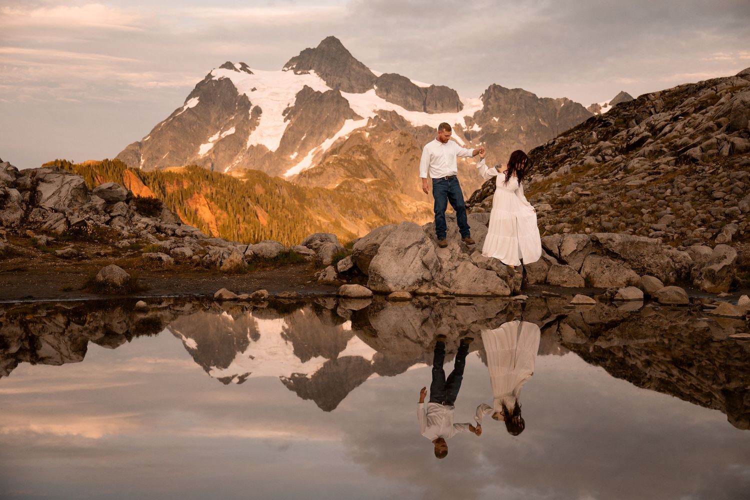 A young couple wanders hand in hand around a small tarn reflecting Mt. Shuksan near North Cascades National Park/Mt. Baker wilderness.