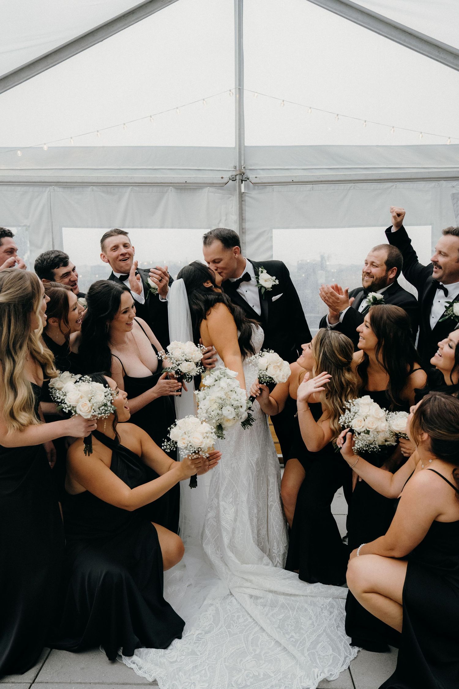 Wedding party in black and white attire celebrates joyfully together in a white tent reception venue.