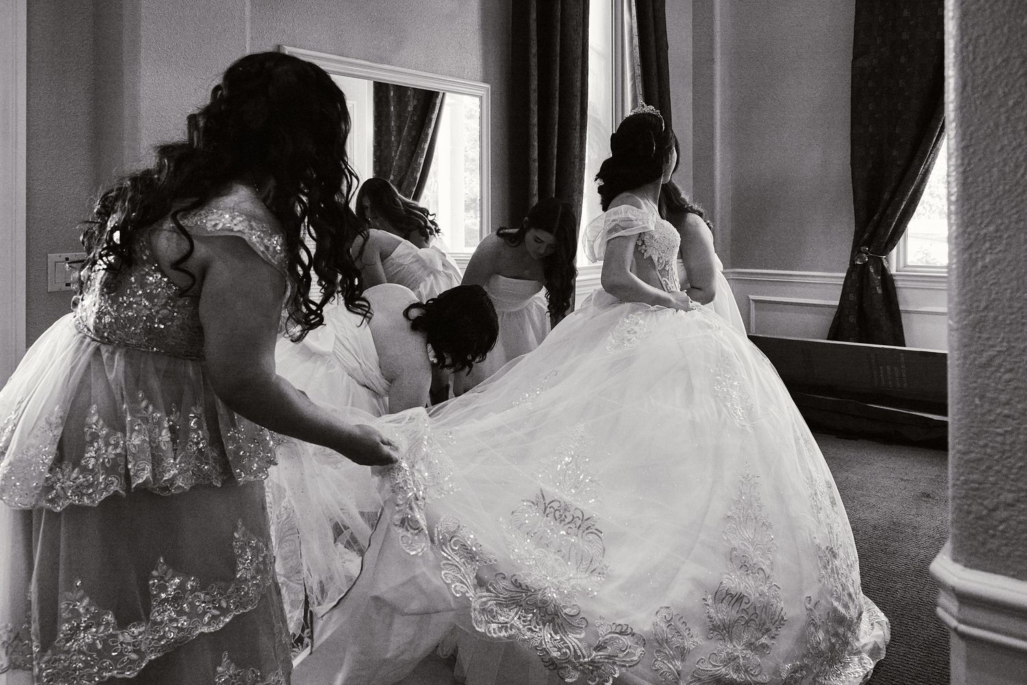 Black and white photo of a bride in a flowing wedding gown being helped with dress adjustments in an elegant room.
