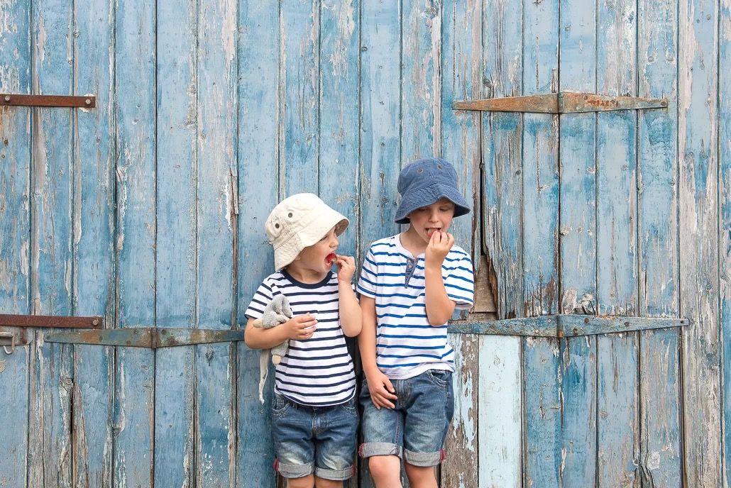 Two children in striped tops sitting and laughing beside a rustic blue wooden wall.