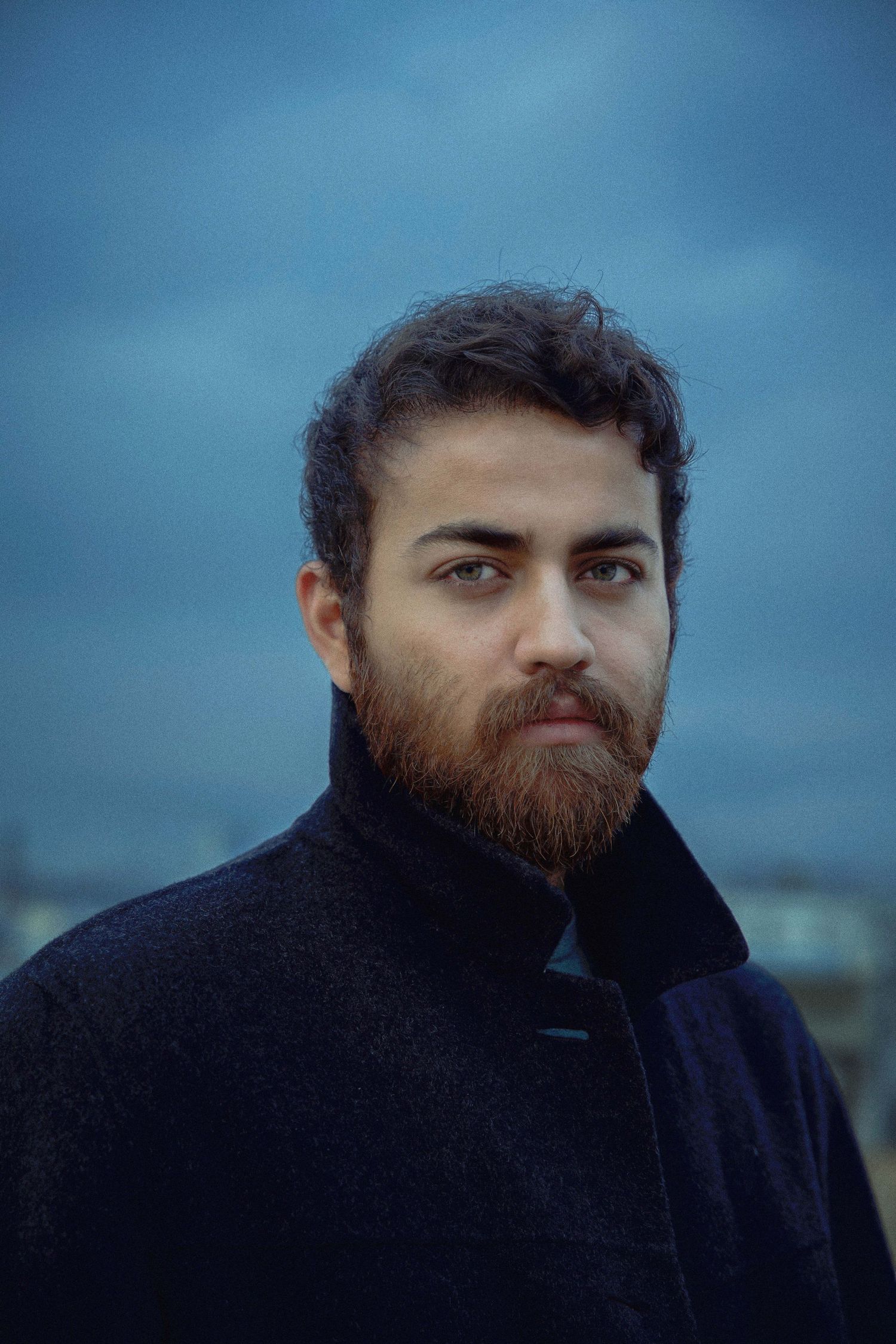 A series of moody portraits showing someone with a dark beard against a blue twilight sky wearing a black coat.