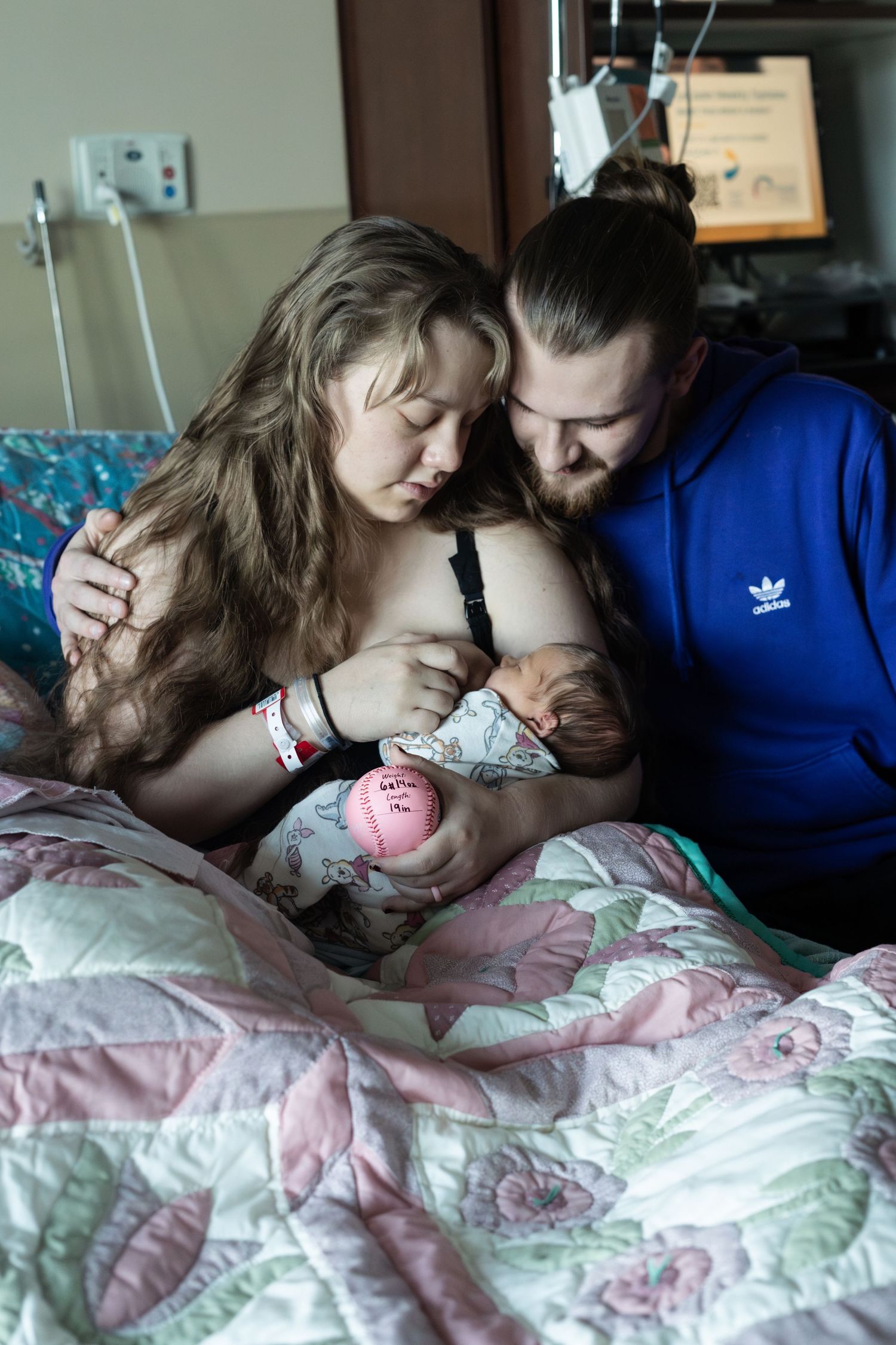 Intimate moment in hospital room with new parents and newborn baby under pink floral blanket.