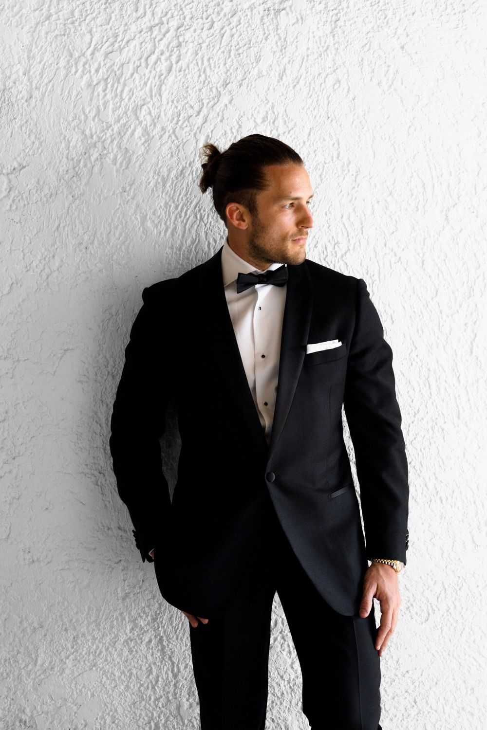 Handsome groom in black tuxedo with bow tie and pocket square leaning against a white textured wall.