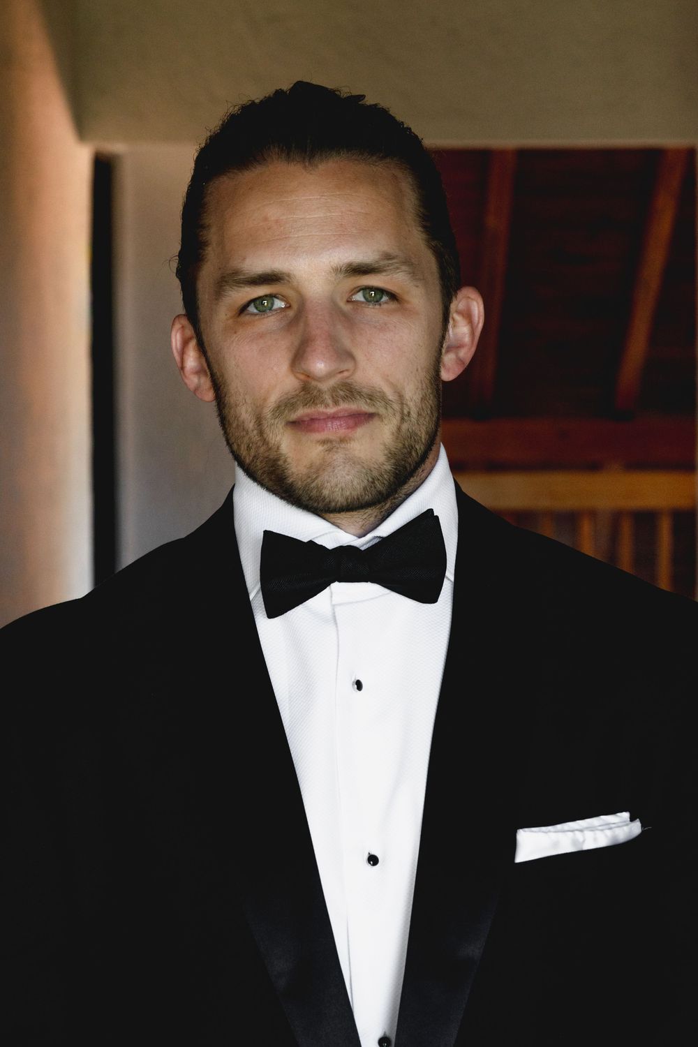A man in a black tuxedo with bow tie and white pocket square poses indoors with a confident smile.