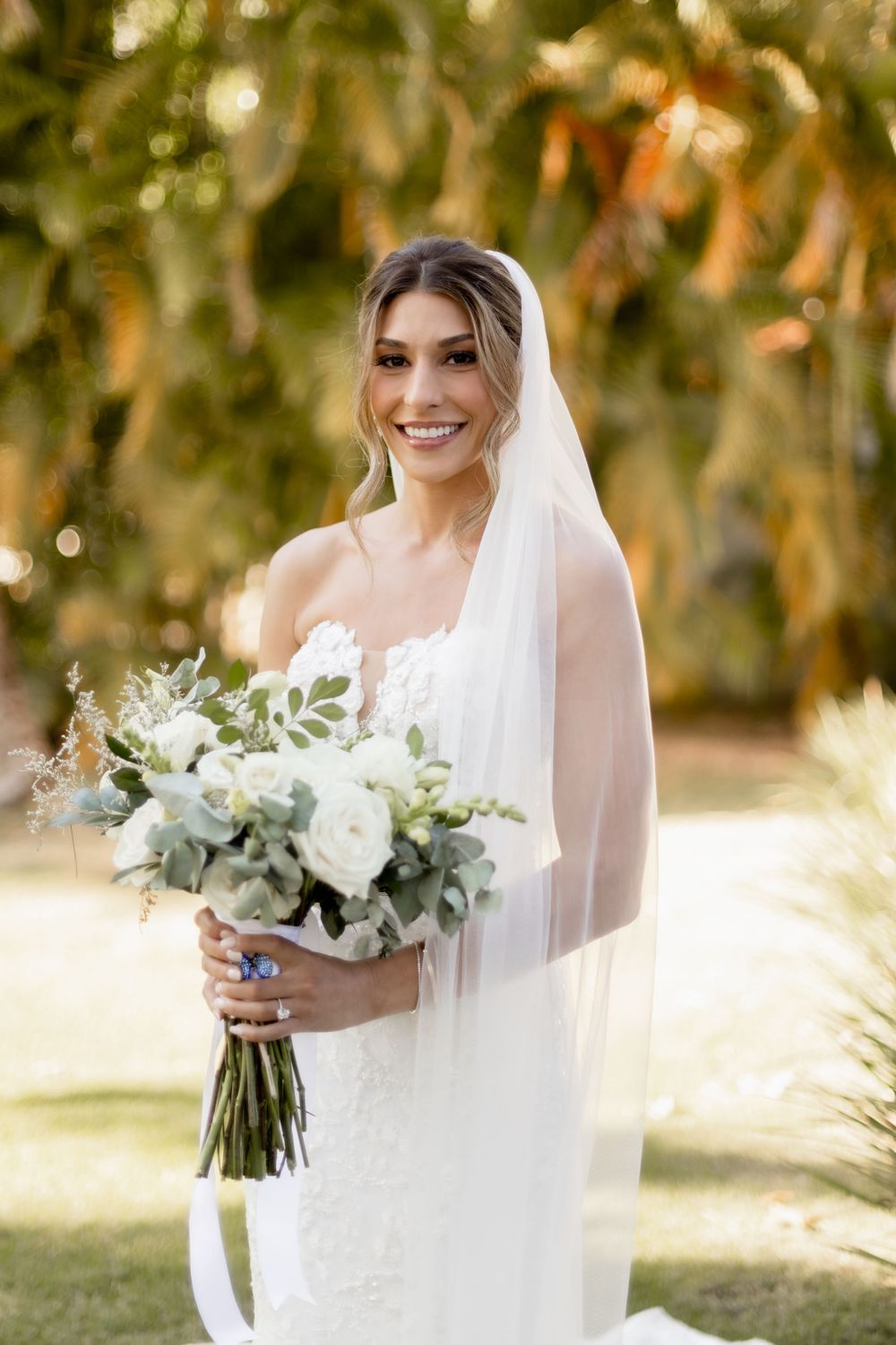 Radiant bride in lace gown holding white and green bouquet with flowing veil outdoors.