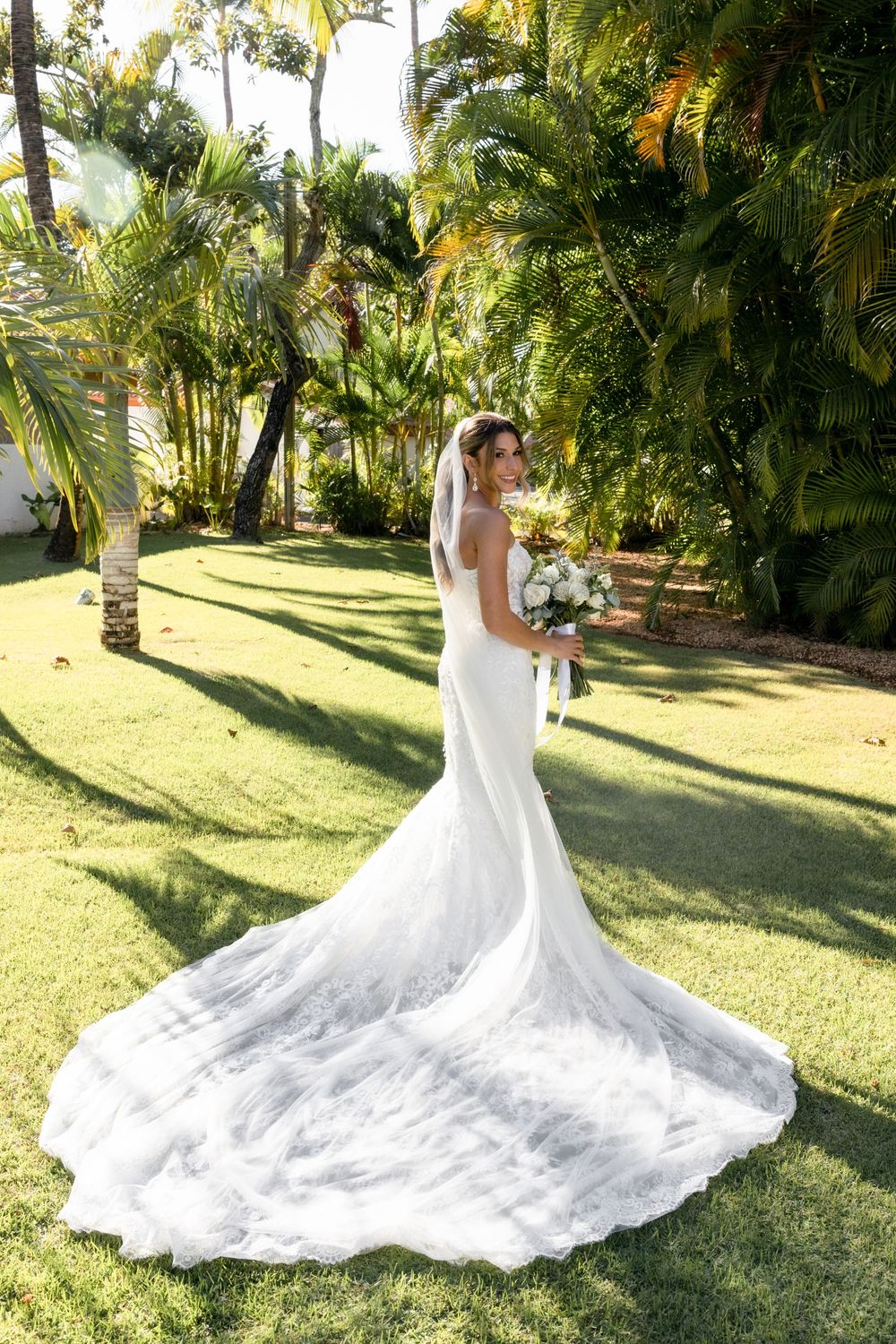 Bride in elegant white gown with long train and veil holds bouquet on lush green lawn surrounded by tropical palms.
