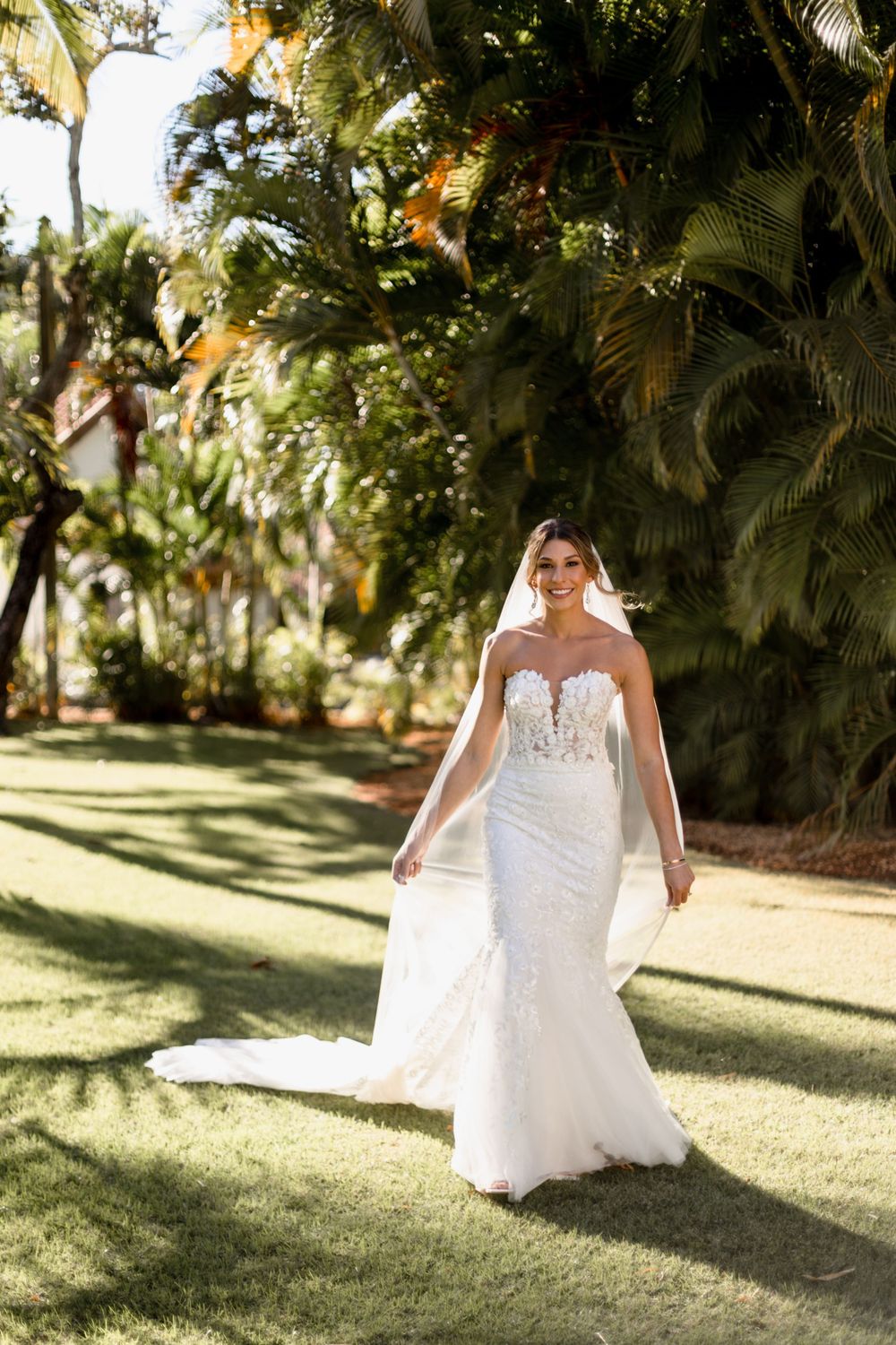 Bride in a strapless lace mermaid wedding gown with veil walks on green lawn surrounded by tropical trees.