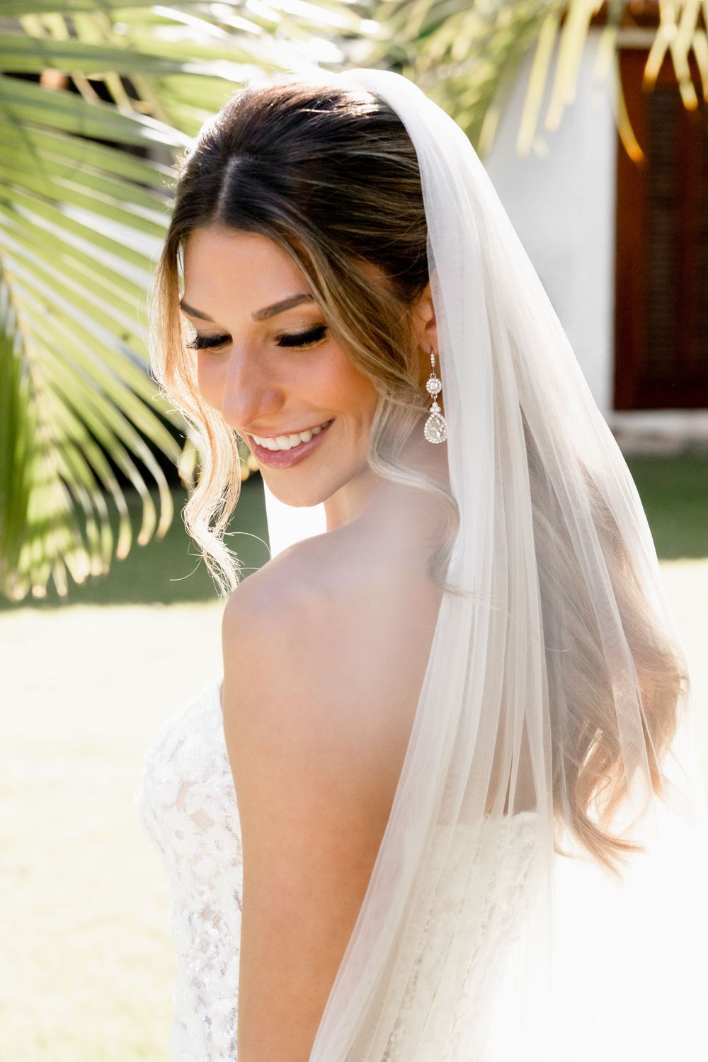 Beautiful bride smiling in white lace gown and veil with drop earrings, standing near tropical palm fronds.