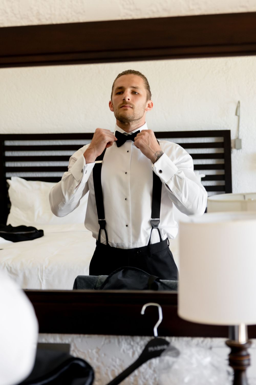 Groom adjusting black bow tie wearing white tuxedo shirt and suspenders in mirror reflection.
