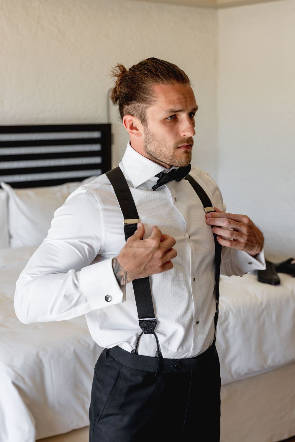 A well-dressed groom adjusting his black bow tie and suspenders over a white dress shirt in a hotel room.