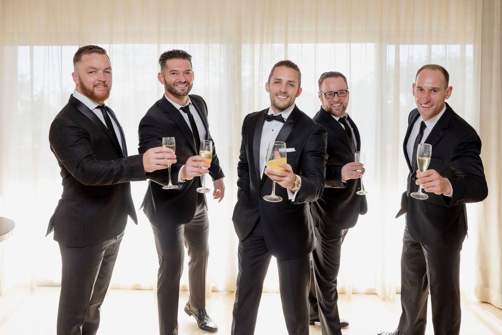 Five groomsmen in black tuxedos toasting with champagne glasses in a bright, curtained room.