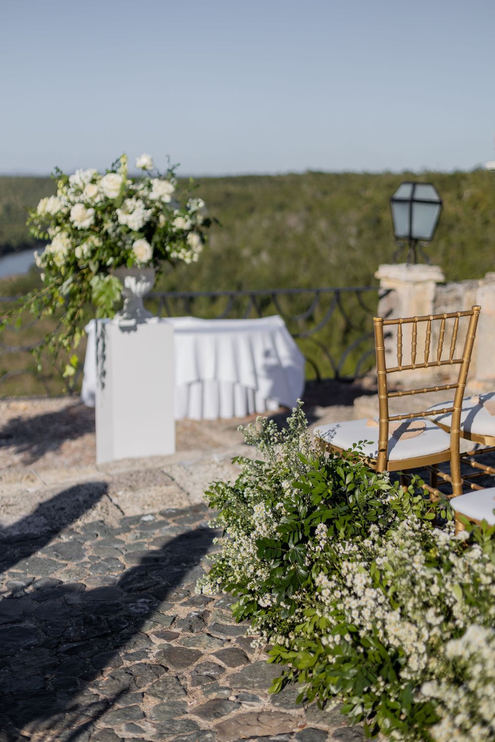 Elegant outdoor wedding setup with white floral arrangements, gold Chiavari chairs, and lush greenery on a stone terrace.