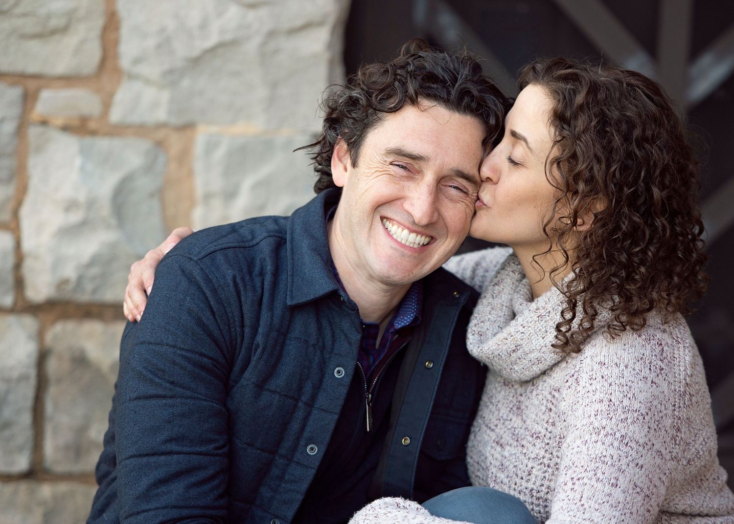 A smiling man receives a kiss on the cheek from a curly-haired woman outdoors near a stone wall.