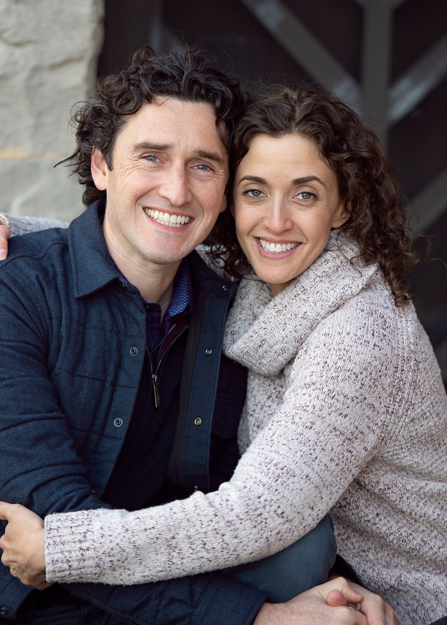 A smiling couple poses closely together outdoors, the man in a navy jacket and woman in a cozy knit sweater.