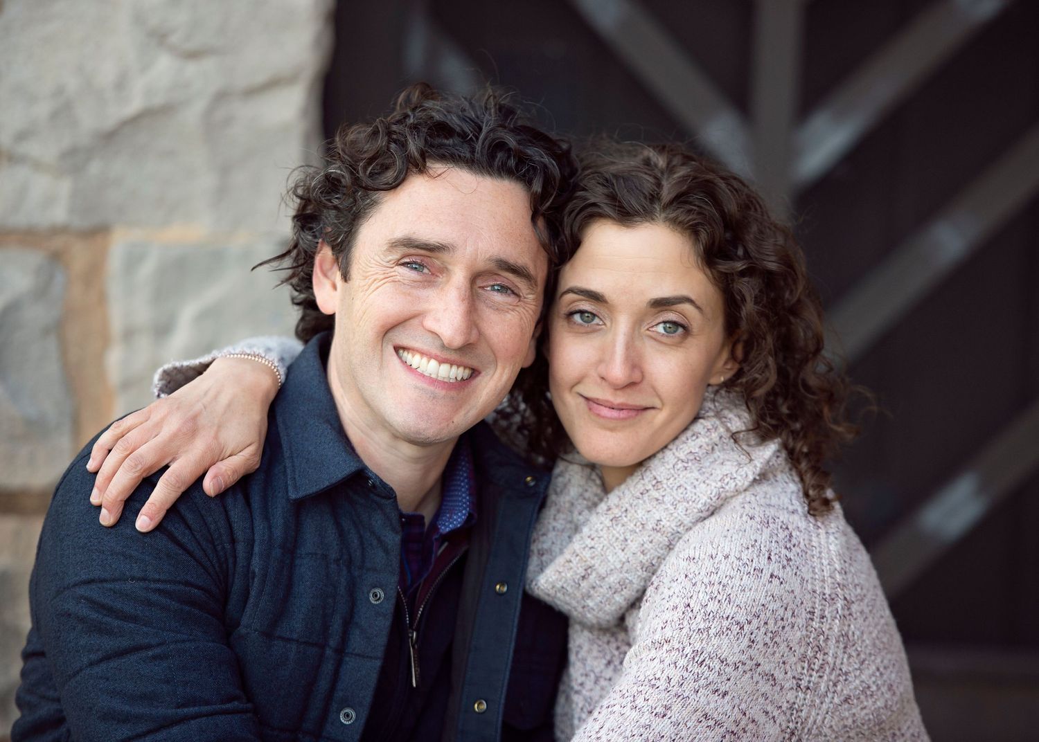 A smiling couple with curly hair poses closely together outdoors, the man in a blue jacket and woman in a cozy knit scarf.