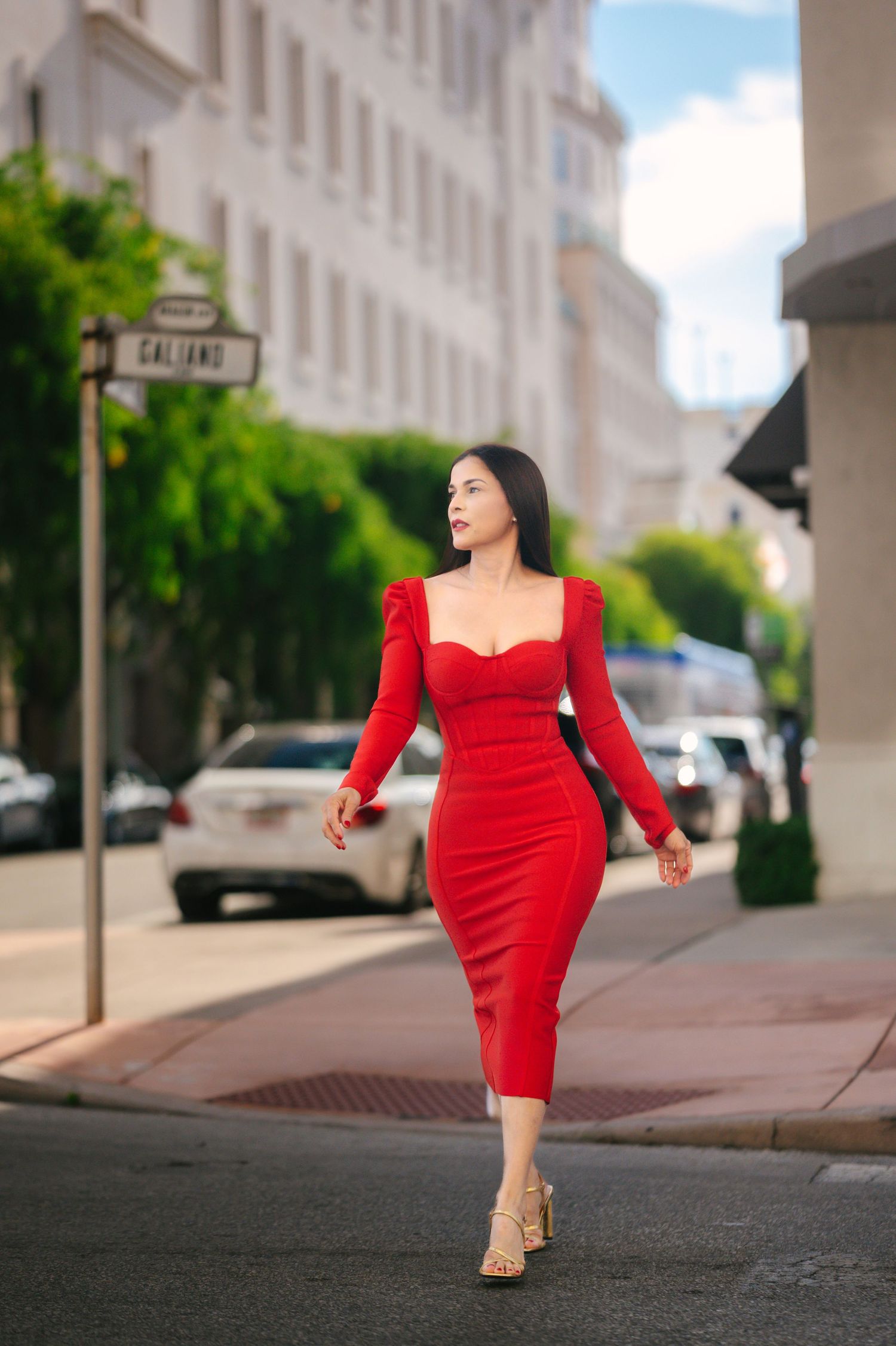 A woman in a vibrant red dress poses confidently on a city sidewalk in front of classic architecture.