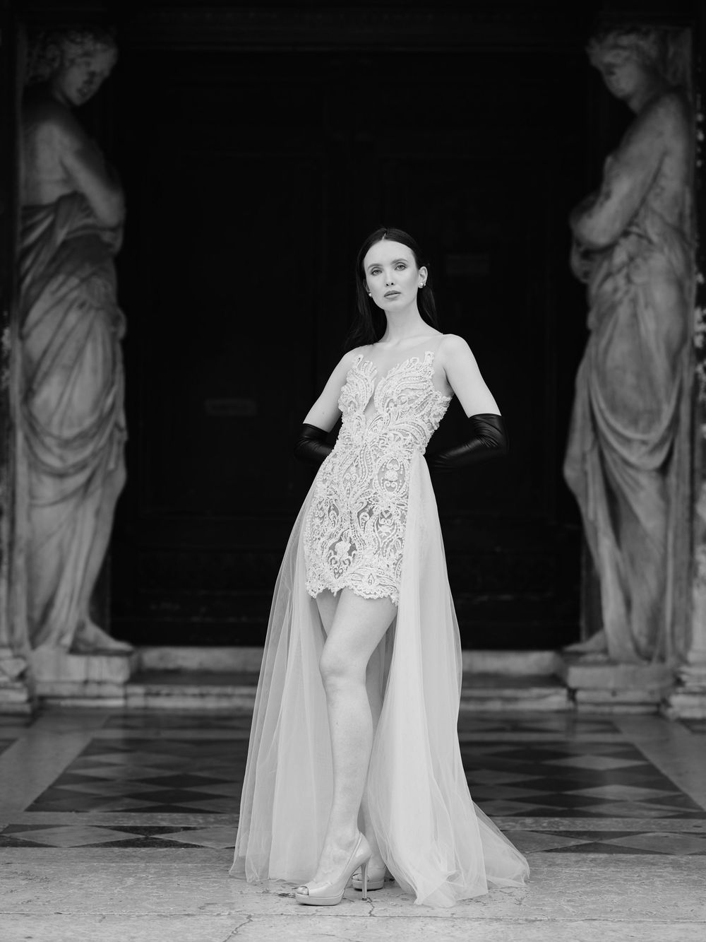 Fashion model walks runway in flowing white gown with lace details and cape during haute couture show.