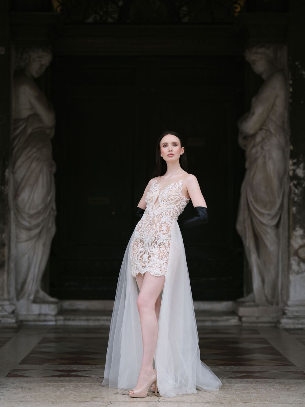 A model walks the runway in a sheer white lace bodysuit with flowing cape at a luxury fashion show.