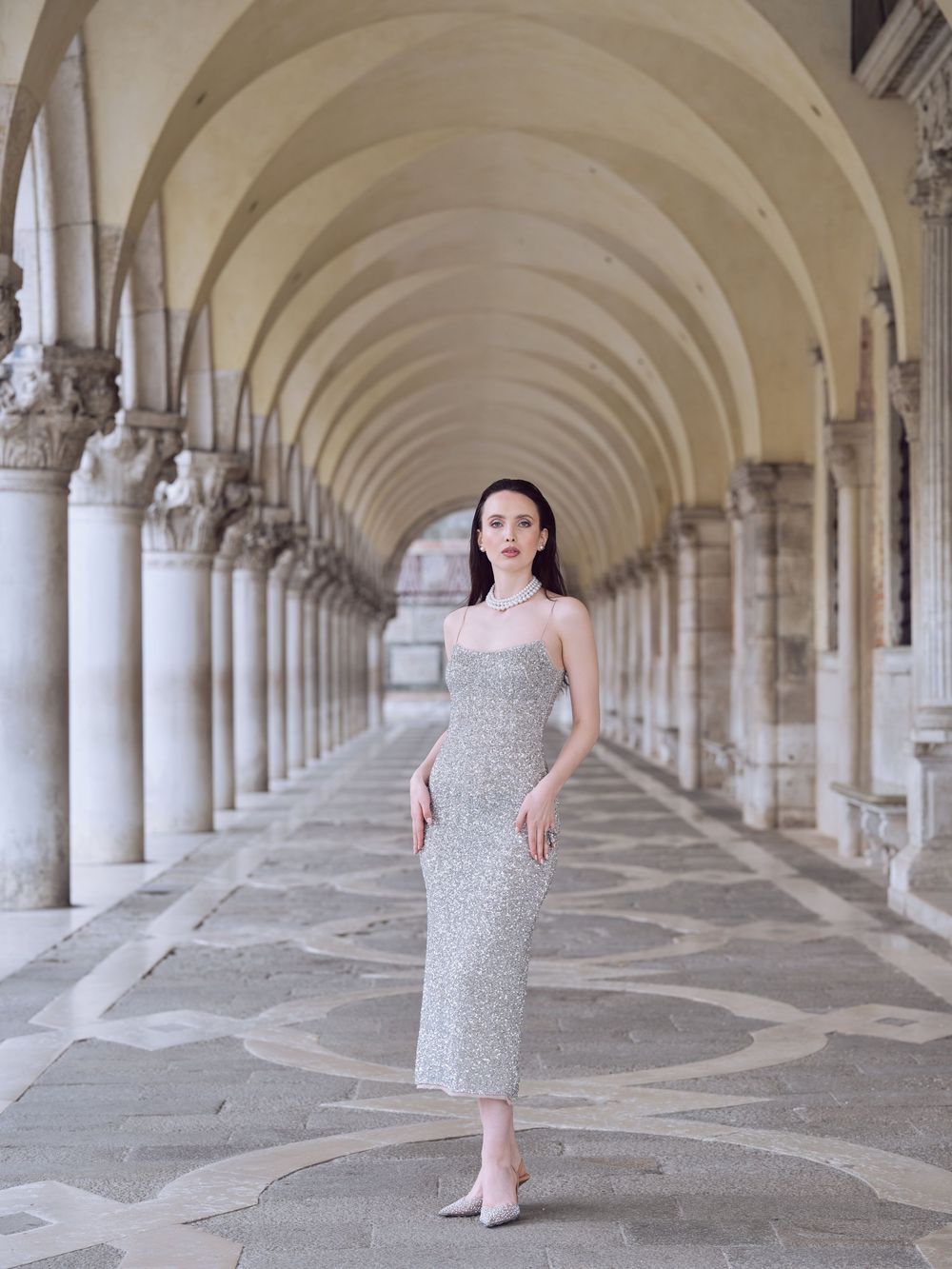 Elegant figure in a silver sequined dress poses in a grand arched colonnade with classical architecture and stone columns.