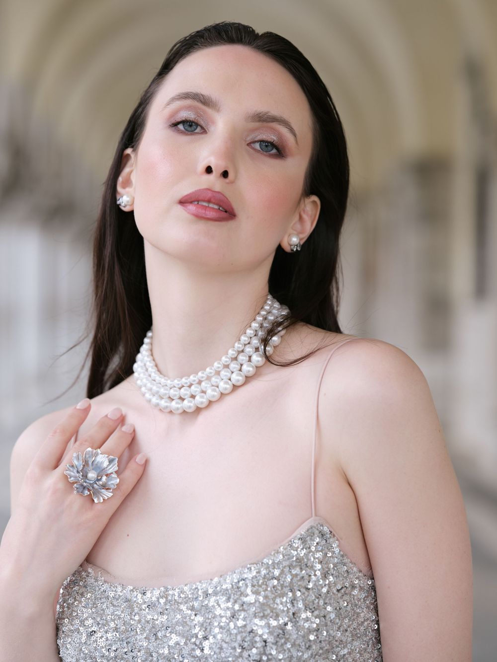 A glamorous portrait in silver sequins and pearls featuring elegant jewelry and dramatic makeup against an architectural backdrop.