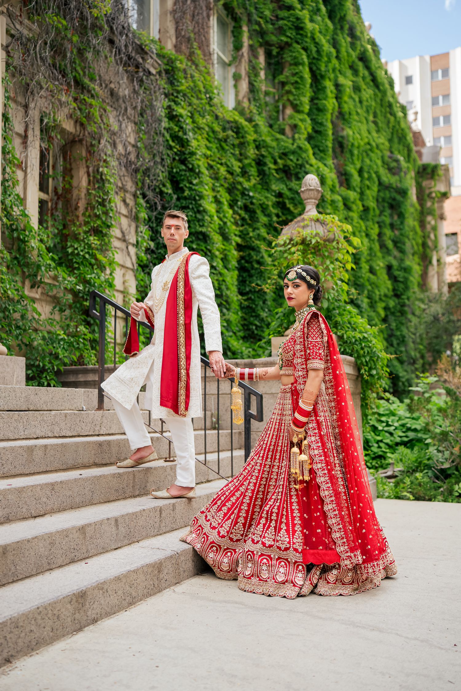 A couple in traditional Indian wedding attire pose on stone steps against an ivy-covered wall.