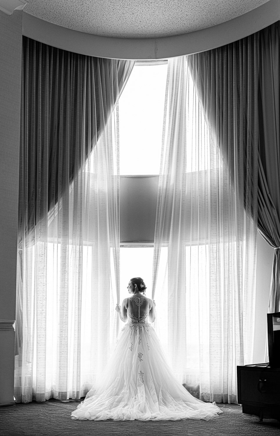 A bride in a flowing white wedding gown stands silhouetted against tall sheer curtains in a dramatic black and white photo.