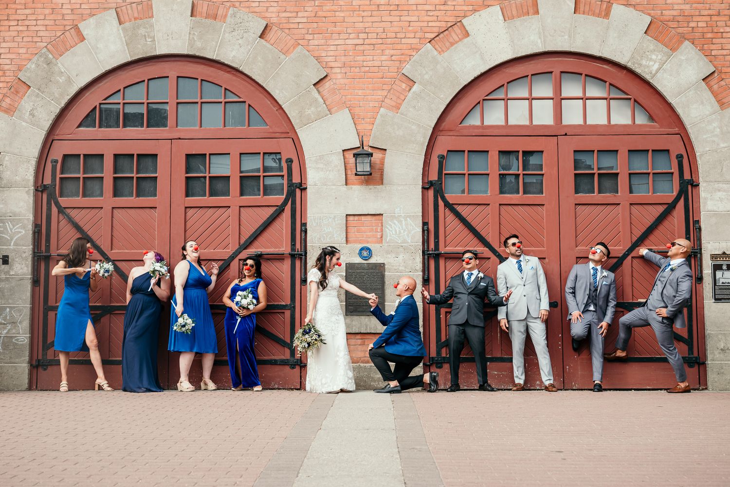 Wedding party in blue dresses poses in sequence against red brick building with arched doorways.