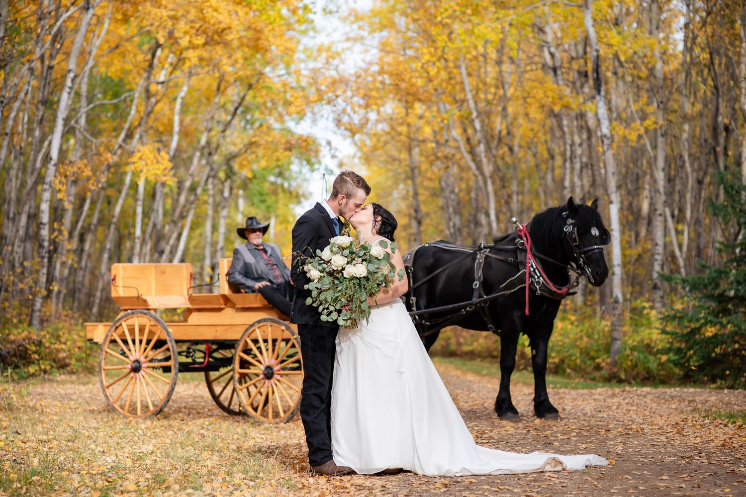 A couple poses in a rustic autumn setting with golden leaves and a wooden wagon in the background.
