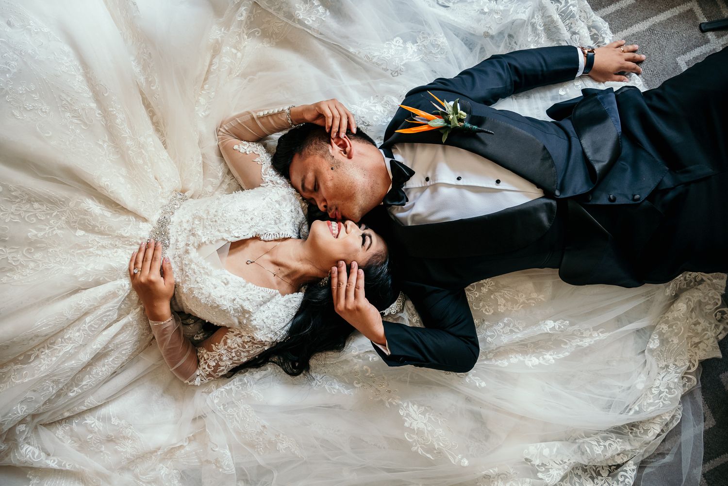 A romantic bride and groom lay side by side on the floor wearing wedding attire in elegant poses.