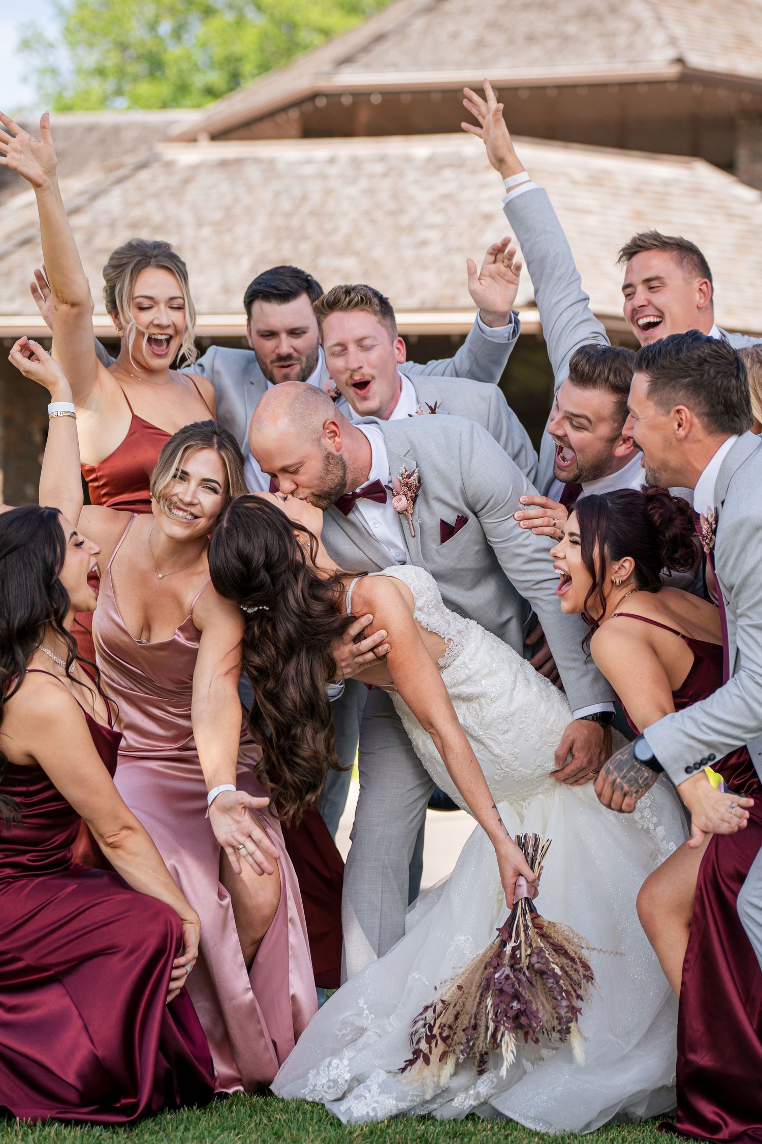 Wedding party in burgundy and gray attire laughs together while posing for candid photo series outdoors.