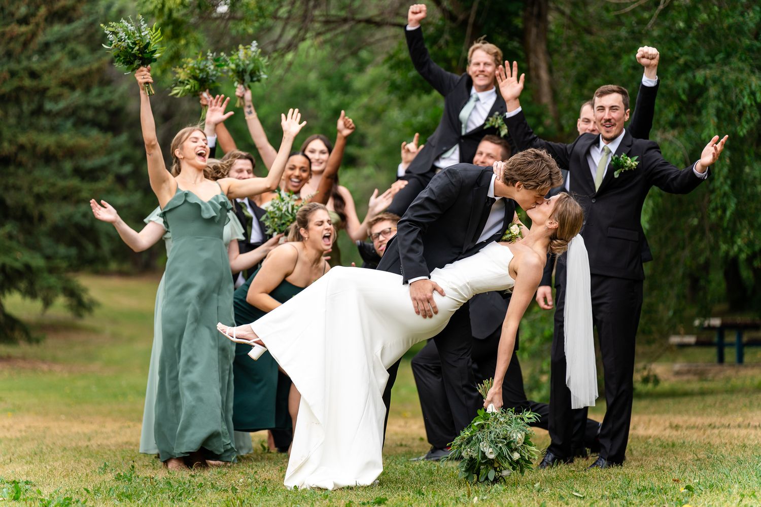 Wedding party poses for fun celebration photo in green garden setting with tuxedos and sage bridesmaid dresses.