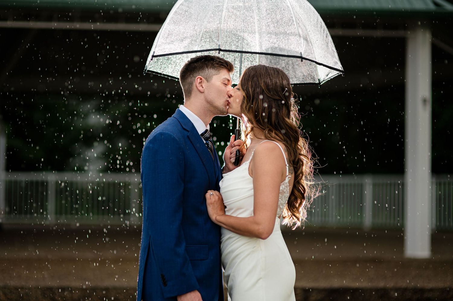 A couple shares an intimate moment under a white umbrella in the rain, the bride in white and groom in navy blue.