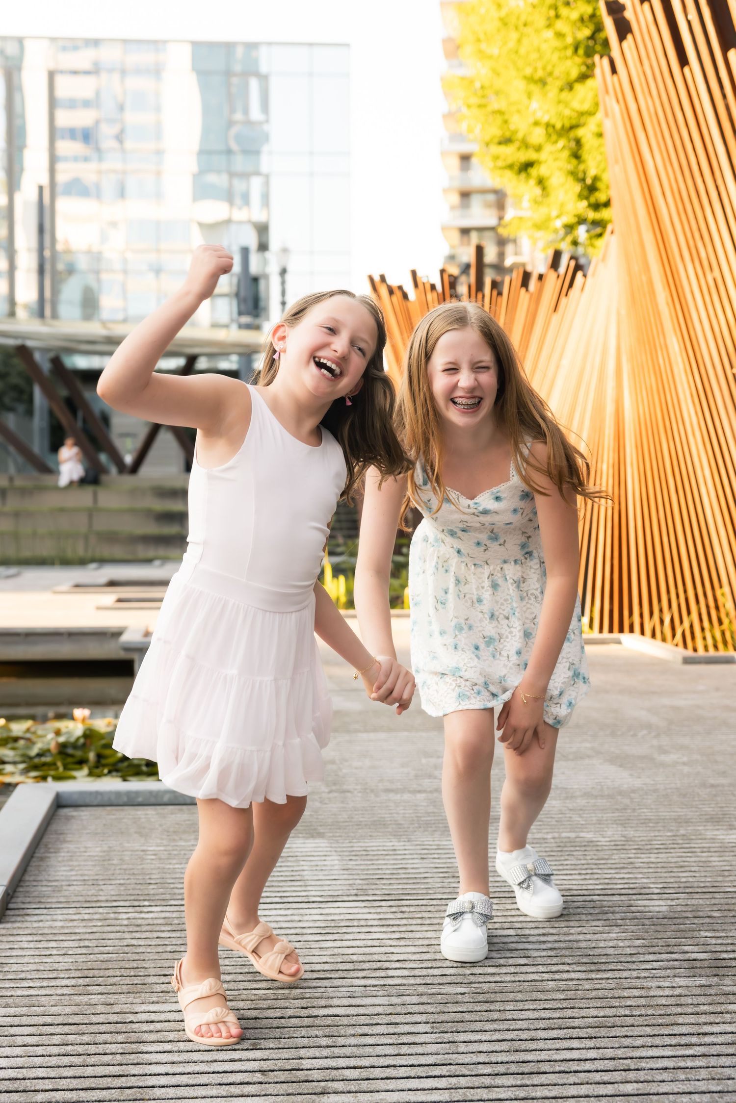Two joyful dancers in light dresses twirl and leap together on a textured outdoor surface in warm sunlight.