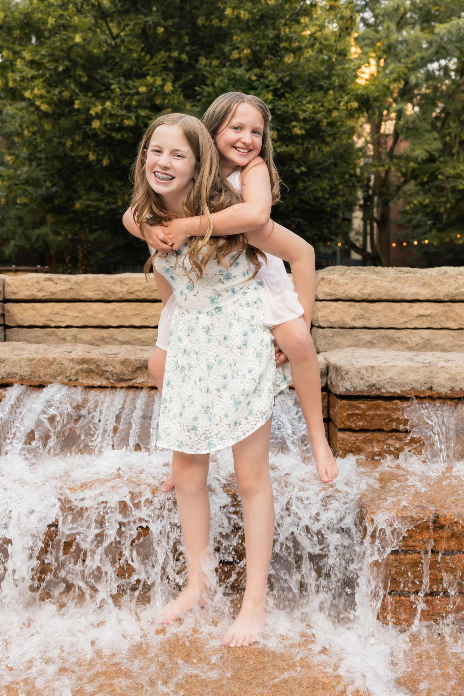 Children in white dresses splash and play by a cascading water feature on a summer day.
