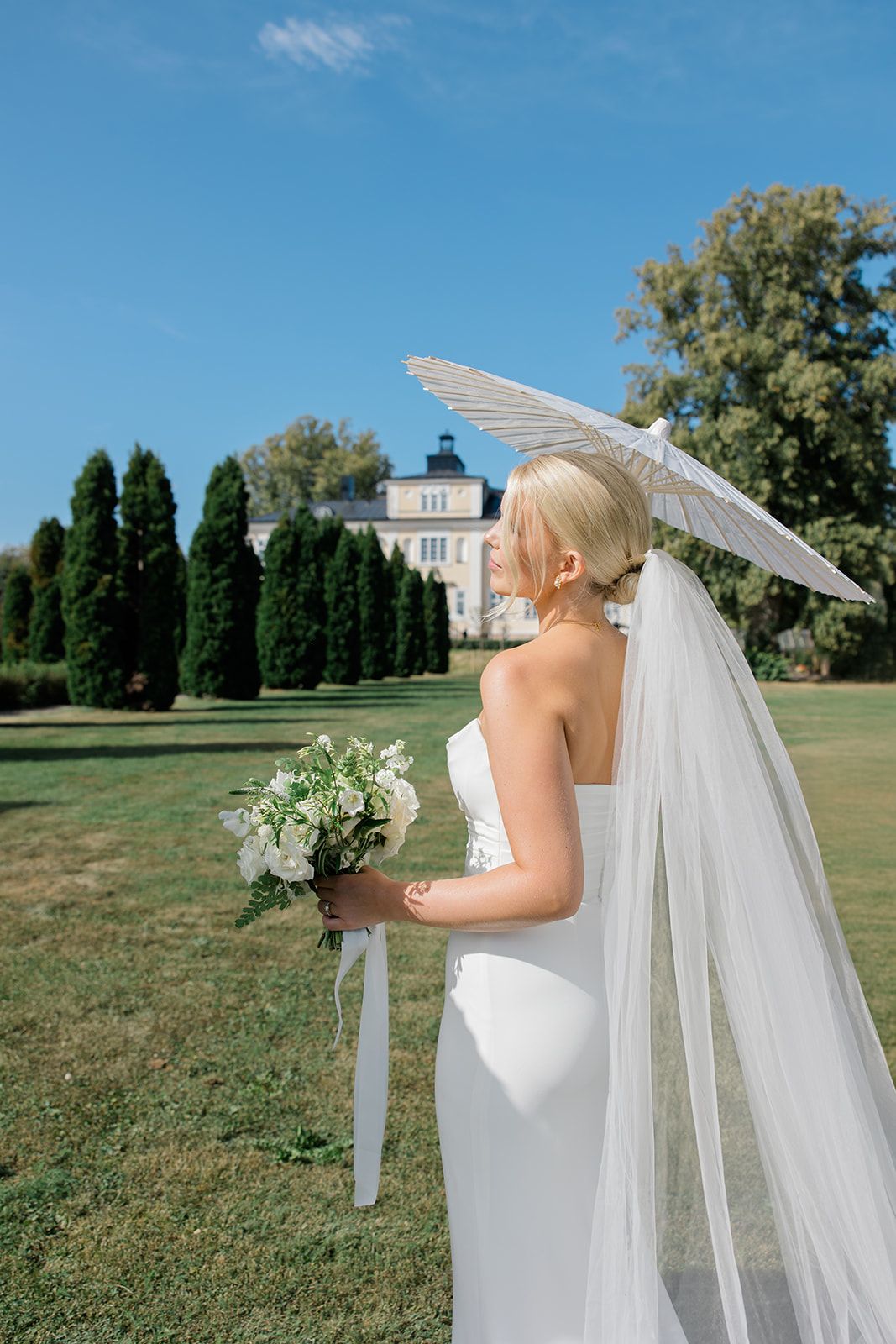 A bride in a white wedding dress and flowing veil stands on manicured grounds with a stately building in the background.