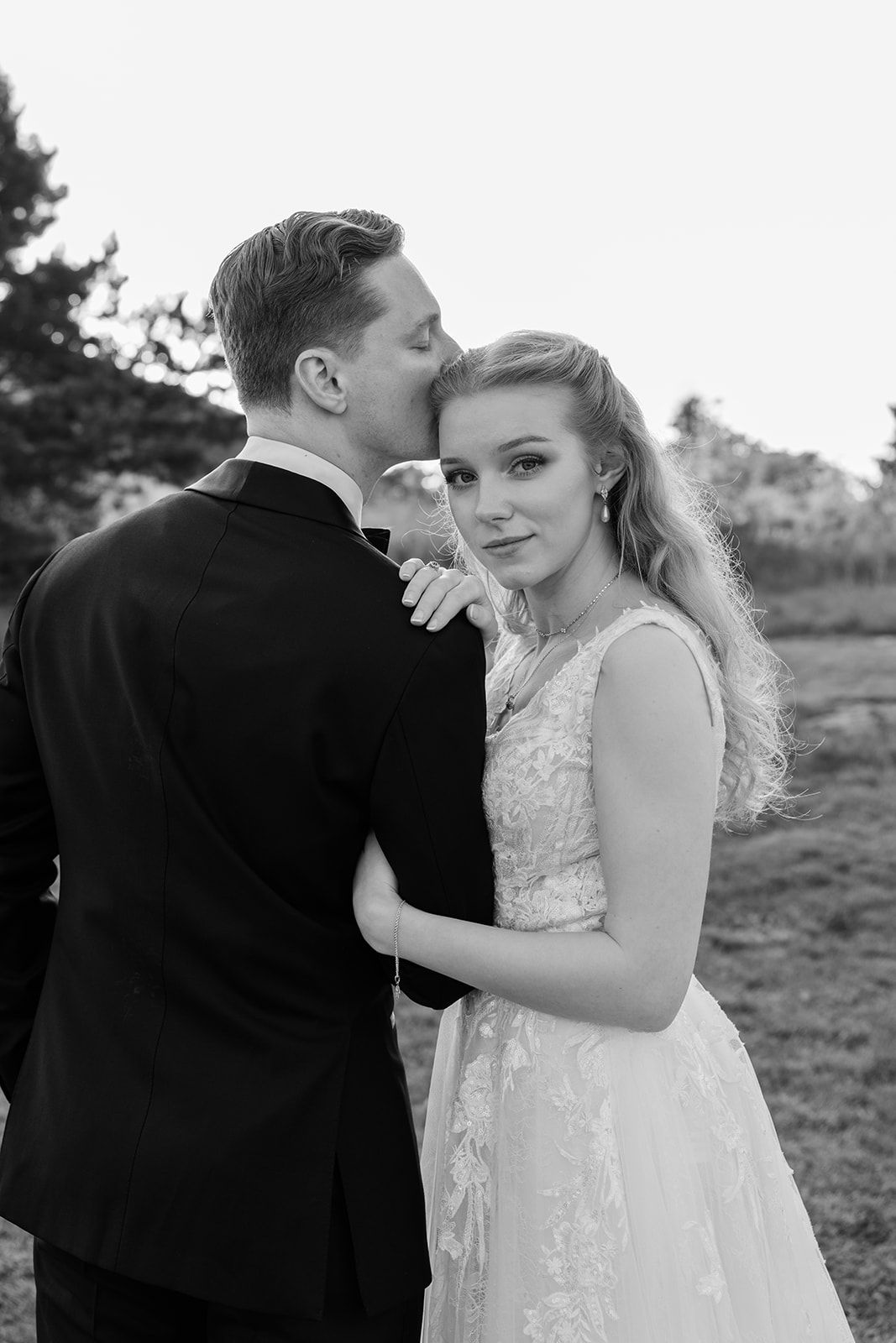 A romantic black and white wedding portrait of a couple embracing outdoors, with the bride in a lace gown.