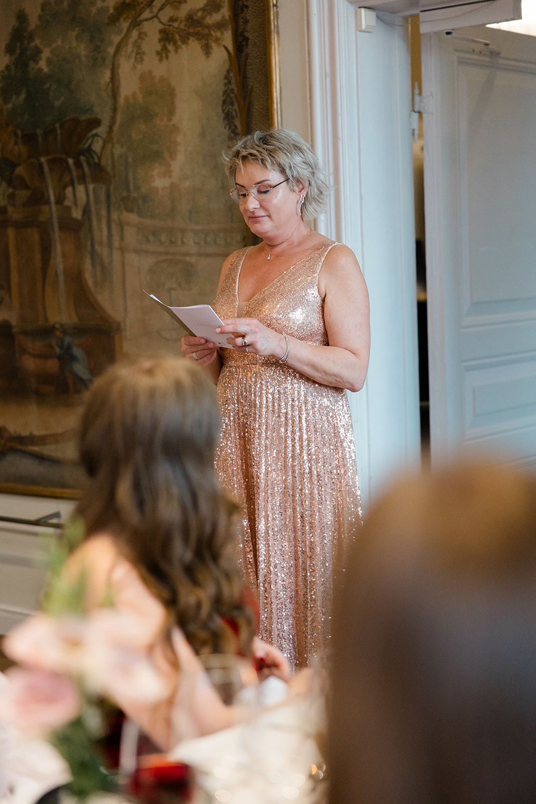 Someone in a sparkly rose gold dress reads from a paper while standing at a podium during a formal event.