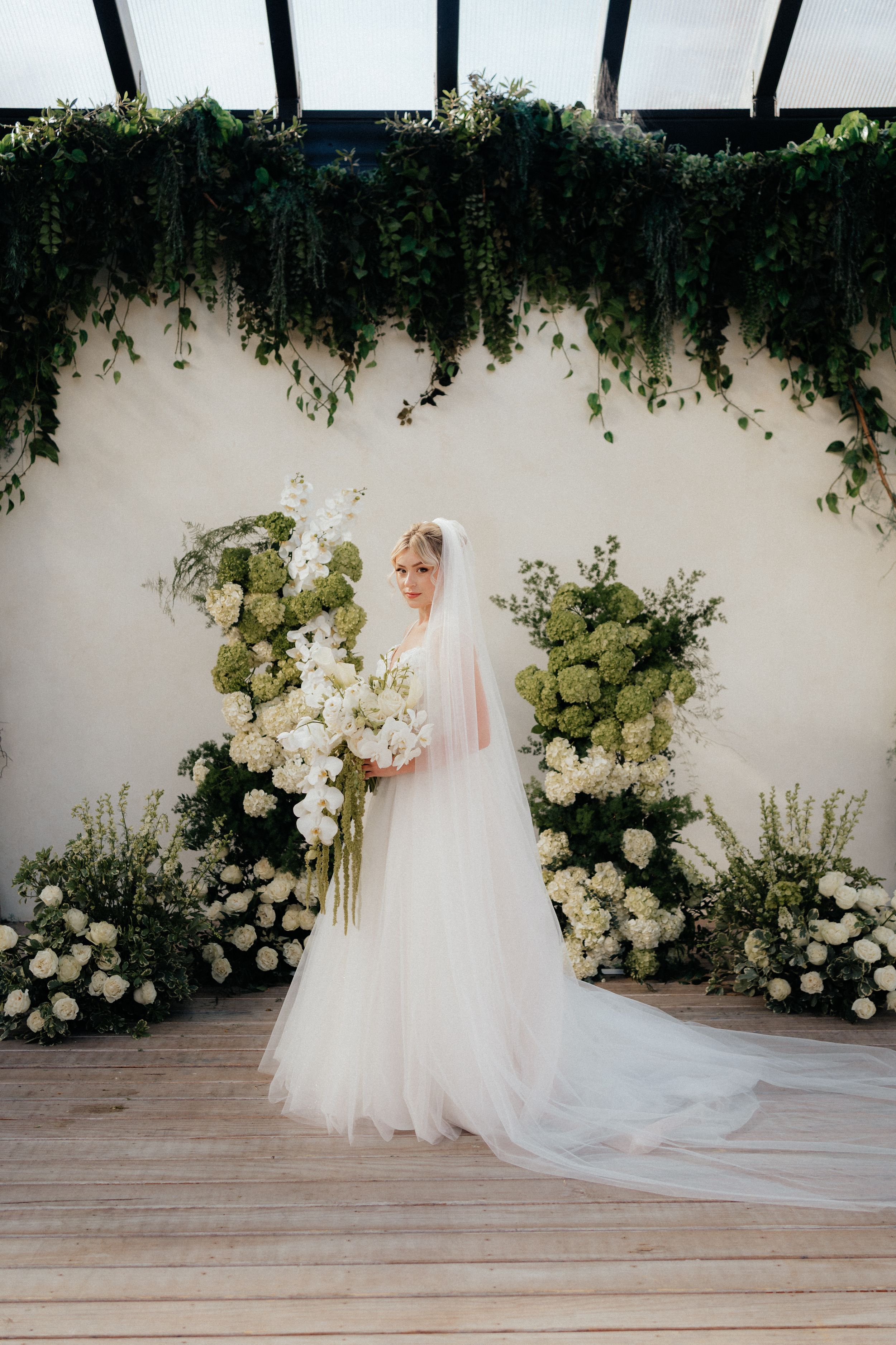 A bride in a flowing white wedding gown and veil poses against a floral backdrop with white roses and greenery.