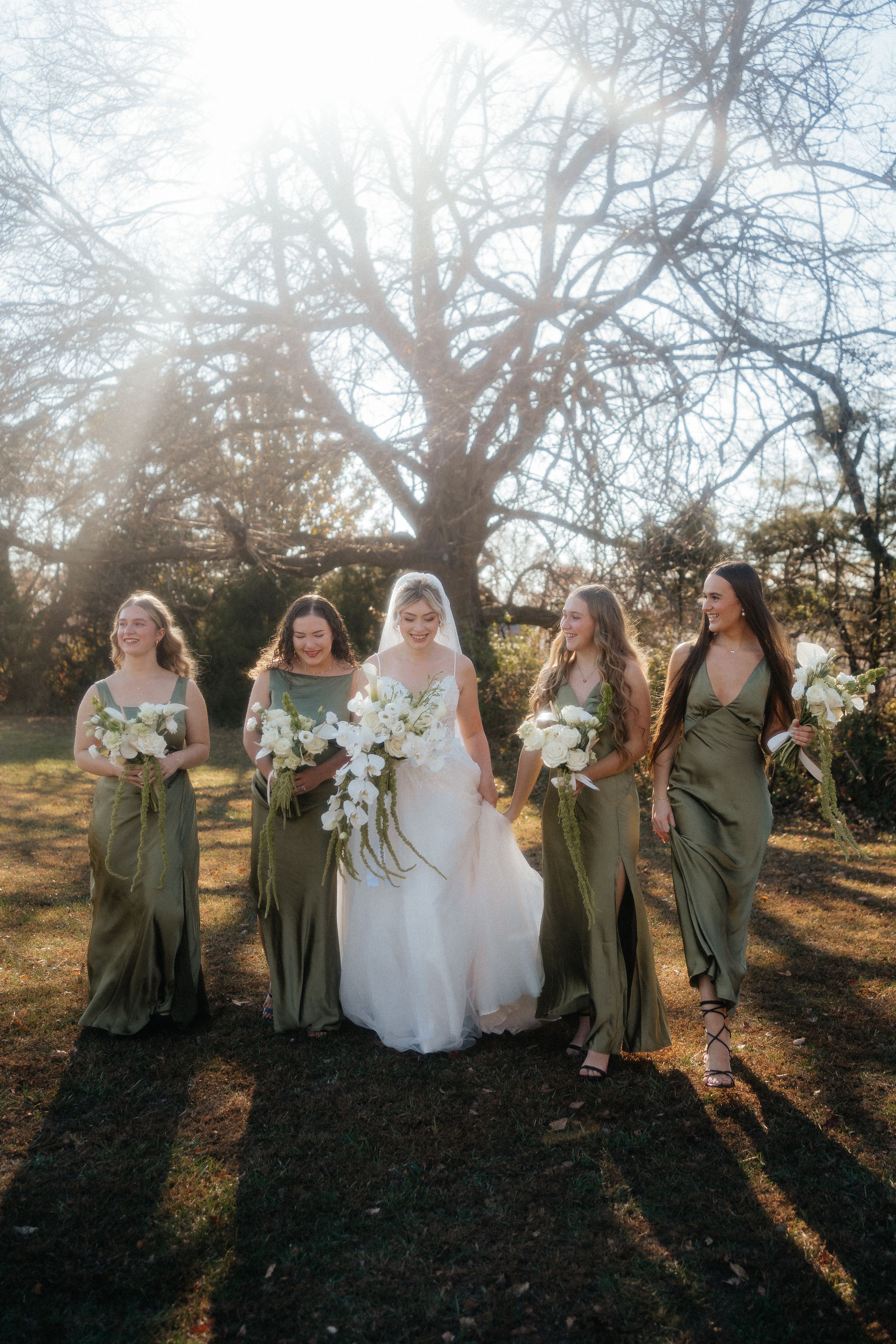 Bridesmaids in olive green dresses pose with bride in white gown against sunlit trees during golden hour.