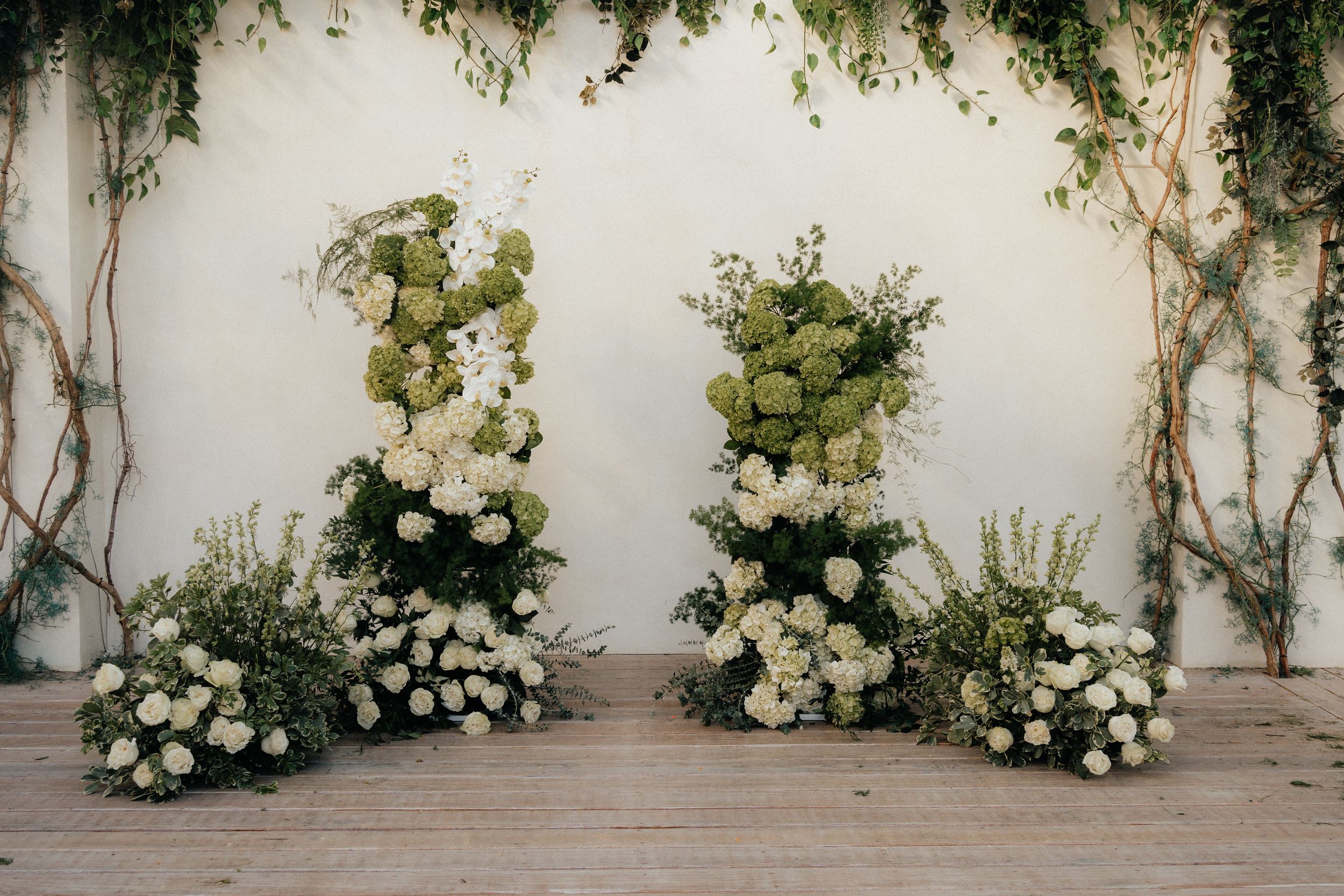 Elegant white floral arrangements and climbing vines decorate a light colored wall at an outdoor event space.