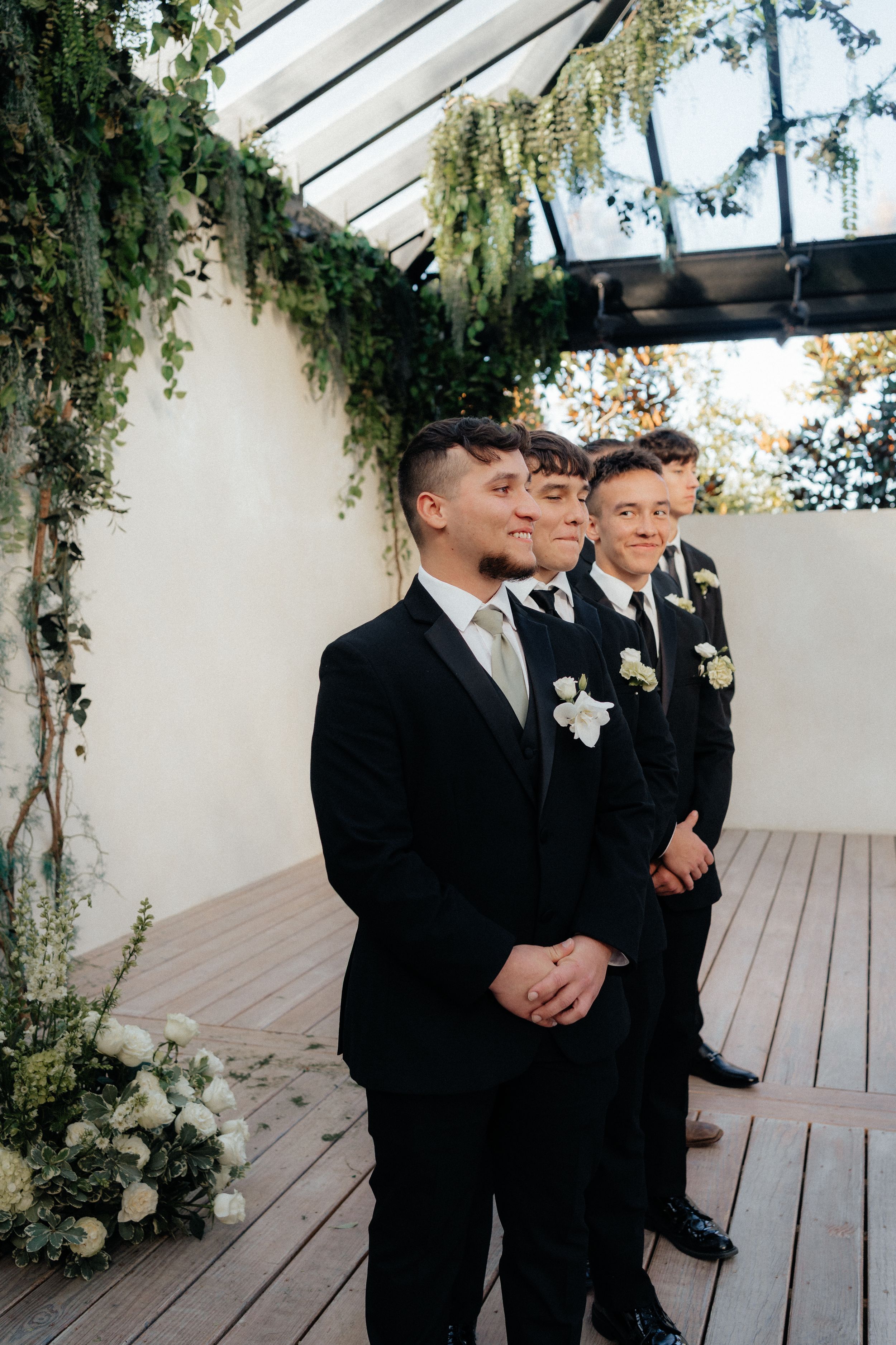A group of groomsmen in black suits stand in a row under hanging greenery at an indoor wedding venue.