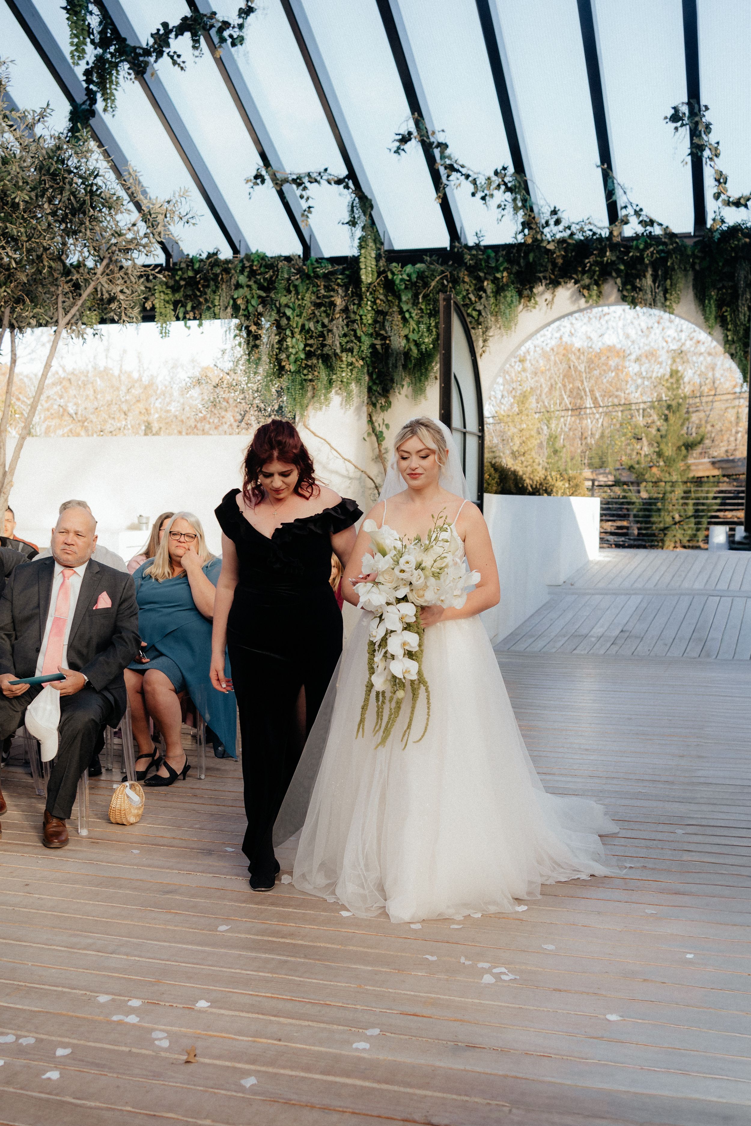 Wedding party performs a fun group dance routine on a wooden deck under a glass-roofed venue.