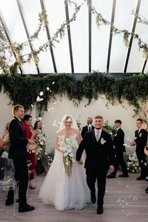 A wedding ceremony takes place in an elegant white venue with floral decorations and guests in formal attire.