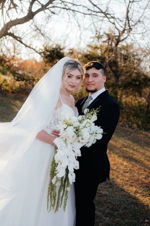 Wedding couple poses together in sunlit outdoor setting with flowing white veil and floral bouquet.