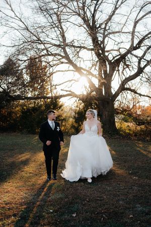 Wedding party walks along a scenic outdoor path at sunset with bare trees casting long shadows.