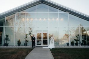 A couple stands outside a modern glass greenhouse venue at sunset during their wedding celebration.