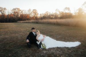 A couple in wedding attire lays together on a grassy field during sunset in a romantic outdoor photoshoot.
