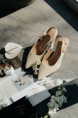 Elegant cream bridal shoes displayed with wedding stationery, white flowers and delicate styling details on concrete surface.