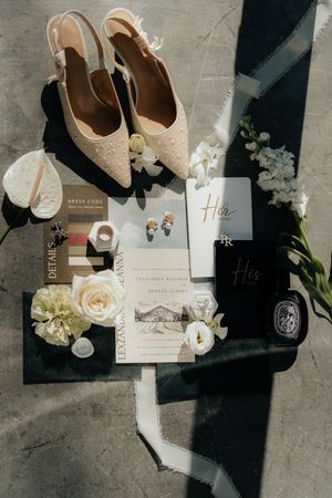 Wedding details flatlay with beige shoes, white floral arrangements, invitations and decor on dark surface.