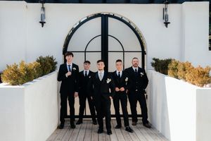 Groomsmen in black suits stand together on white porch with arched architectural details.